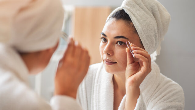 A beautiful young woman plucking her eyebrows in the mirrror