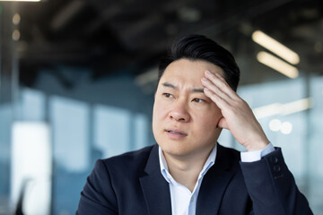 Close-up photo of a young Asian man in a suit sitting at a desk in the office and holding his head,...
