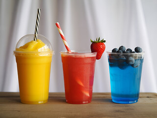 Three colorful drinks in plastic cups with straws and fruit garnishes on a wooden surface view