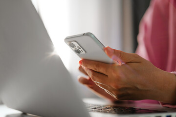 Close-up of hands holding a smartphone while working on a laptop, with warm light filtering in from the background.