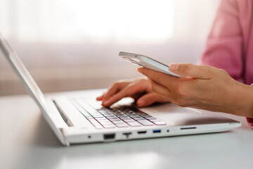 Fototapeta premium Hands type on a laptop keyboard while holding a smartphone, showcasing multitasking in a modern work environment.