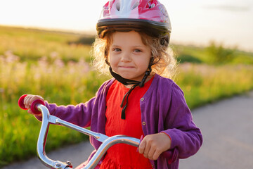 Obraz premium A cute little girl in a pink helmet and a purple cardigan looks directly at the camera while holding her bicycle handlebars, with a sunlit grassy field in the background.