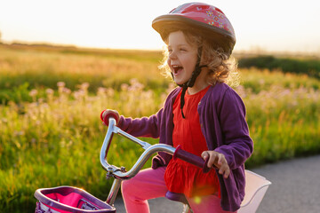 A joyous little girl in a helmet laughs exuberantly while sitting on her bicycle against a golden,...