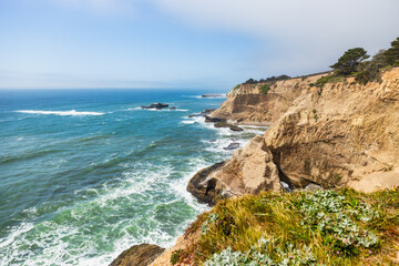 Waves crash against sculpted sandstone cliffs and sea caves along the rugged Pacific Coast near Highway 1 in California. A small hidden beach nestles at the base of the cliffs
