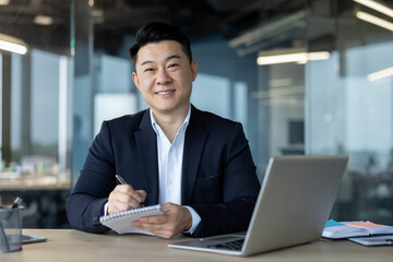 Portrait of a young Asian man working in an office, sitting at a desk, working on a laptop and...
