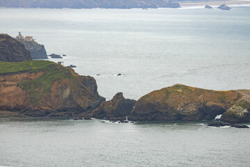 Coastal rock formations and the iconic Point Bonita Lighthouse seen from afar in the Marin Headlands, with waves crashing gently against the rugged shoreline