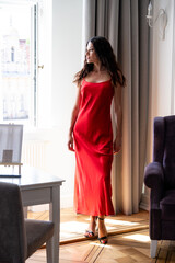 Full-length portrait of stylish brunette model in long red gown walking upstairs and posing near...