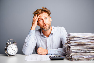 A stressed man sits at a desk with a large stack of paperwork, a calculator, and a clock, looking overwhelmed by his workload.