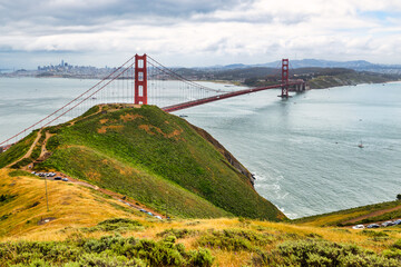 Panoramic view of the Golden Gate Bridge spanning San Francisco Bay with cityscape, lush hills, and boats, captured from a high vantage point on a cloudy spring day