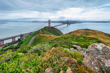 Panoramic view of the Golden Gate Bridge spanning San Francisco Bay with cityscape, lush hills, and boats, captured from a high vantage point on a cloudy spring day
