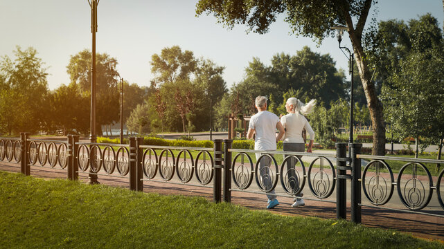 Rare view of sporty senior couple in sportswear jogging along a bridge in park having sport workout at sunset. Elderly man and woman running outdoors. Jog workout in nature for retired people concept.