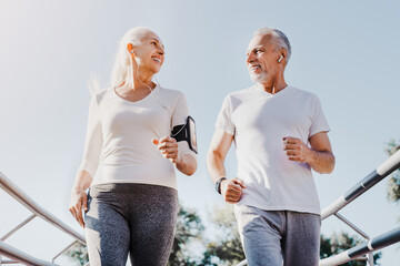 Cheerful active senior couple looking to each other with love and smiling jogging together doing...
