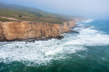 Aerial view of Californias rugged coastline along Highway 1, with steep cliffs, turquoise waters, and sea caves under a bright sky, capturing the wild beauty of the Pacific shore