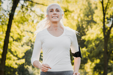 Front view of senior woman running in a park with a smile on her face and earphones on. Elderly...