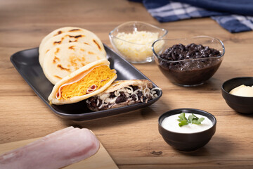 Close-up of Venezuelan arepas, one filled with shredded cheddar and ham, the other with black beans and white cheese. Latin American breakfast or lunch dish. Companion ingredients are on the side.