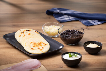 Traditional Venezuelan arepas on a plate with black beans, shredded cheese, ham, sour cream, and butter on the side, ready to be filled for breakfast or lunch.