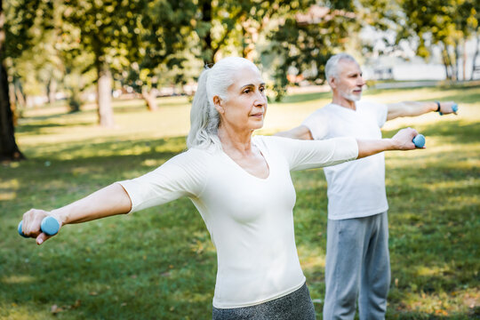 On a sunny day in a vibrant park, a cheerful mature couple lifts dumbbells together, showing their commitment to fitness and healthy aging through fun outdoor exercises accessible to the community
