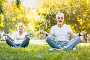 Yoga practice at park. Peaceful grey haired man and woman sitting in lotus pose with closed eyes and meditating on fresh air. Couple on retirement keeping hands in mudra gesture and feeling calmness.