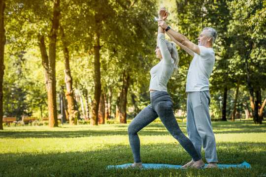 Senior fitness instructor trainer coach helping elderly woman stretching warming up during an outdoor workout session. They smiling and enjoying exercising together. Rehabilitations yoga class concept