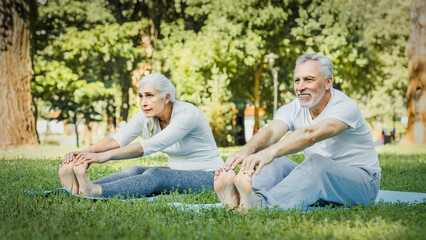 Flexible exercises for body. Sporty man and woman with grey hair stretching on yoga mats with hands to one leg during outdoors workout. Happy married couple with bare feet warming up together at park.