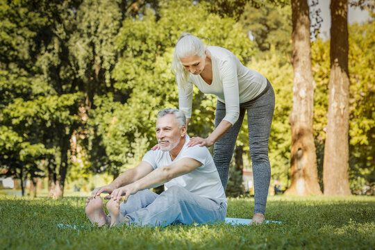 Senior fitness instructor is helping an elderly man stretching his legs during an outdoor workout session. They are both smiling and enjoying the benefits of exercising together - Powered by Adobe