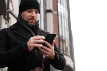 Handsome man wearing black coat and brown leather bag walking down on the city with mobile phone in...