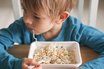 6 years old boy eats instant noodle from box. Child is eating spaghetti. Lunch time. A good appetite. Tasty children food. Close-up portrait. Carbs dinner. Fast food. Kid eating by himself with fork.