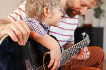 Handsome dad teaches his cute son to play the guitar sitting on sofa in the living room at home....