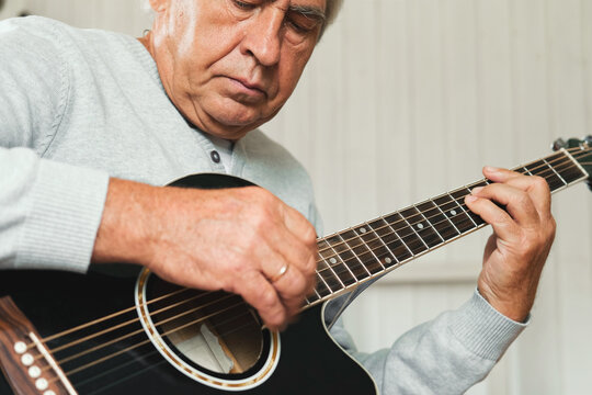 Senior man is playing guitar. Elderly male sitting at sofa and play music. Portrait of a gray haired mature 60s person with acoustic guitar, he is learning to play. Enjoying retirement life at home.