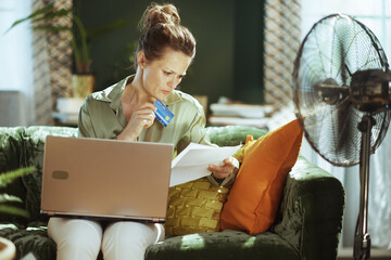 A focused woman manages home finances, reviewing documents with a credit card & laptop. Seated on a green velvet sofa, she navigates bill payments; a floor fan hints at the warm indoor environment.