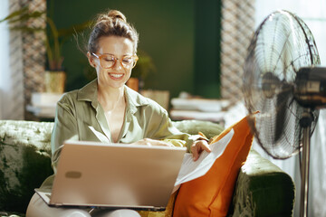 A smiling woman with glasses works comfortably from her green sofa, holding papers and using a laptop. A floor fan nearby provides relief, highlighting a productive home office setting in warm weather
