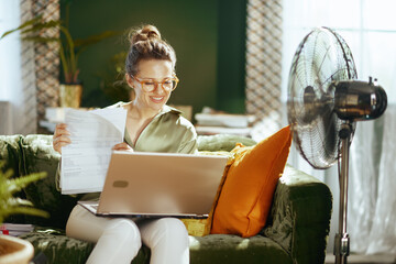 A smiling woman with stylish glasses and a messy bun sits comfortably on a green sofa, working on her laptop. A fan in the background suggests a warm, productive summer day in her home office.