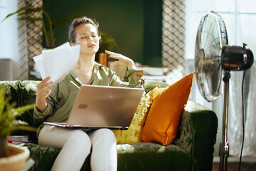 A middle-aged woman on a green velvet couch, fanning herself with papers, shows discomfort. A fan nearby emphasizes her struggle with hot indoor conditions while working.