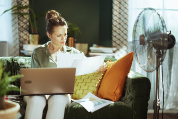 A dedicated middle-aged woman intently reviews documents and charts on her green velvet couch, with a laptop nearby. Depicts focused financial management or business work from a home office.