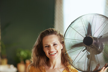 A cheerful adult woman with long wavy hair smiles confidently while holding a large metal floor fan. This bright indoor setting evokes comfort and relief from summer heat at home.