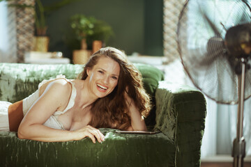 An adult woman on a green velvet sofa laughs joyfully at the camera, embodying comfort & relaxation. With a tablet & a fan cooling the bright indoor space, its a scene of pure contentment.
