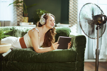 A young adult woman, smiling brightly, lies on a green velvet couch with over-ear headphones, engrossed in her tablet. A standing fan completes this relaxed scene of joyful home entertainment.