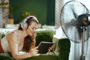 A smiling woman lies comfortably on a green velvet sofa, engaging with her tablet through headphones. A standing fan and blinds indicate a cooled indoor space for leisure and digital enjoyment.