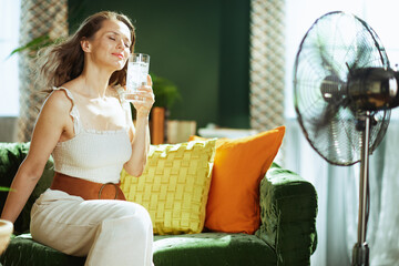 A content woman with eyes closed relaxes on a green velvet sofa, enjoying a glass of iced water and the breeze from a floor fan, finding comfort and relief from the summer heat indoors.