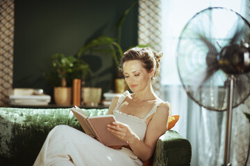 An adult woman enjoys a quiet moment, comfortably reading a book on a green velvet sofa in her sunlit living room, with a standing fan nearby for comfort.