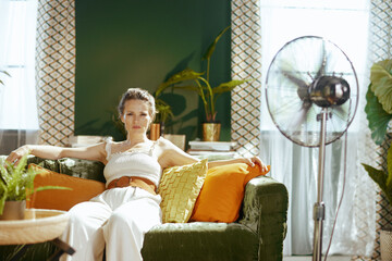 A relaxed woman in a crop top & wide-leg pants sits on a green velvet sofa, smiling at the camera. A standing fan in her bright, plant-filled living room emphasizes a comfortable, cool domestic scene.