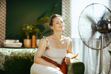A serene woman finds comfort and relief on a green velvet sofa, eyes closed as she basks in the cooling breeze from a standing fan in her brightly lit, modern living room.