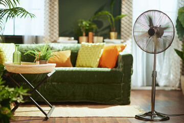 A bright & stylish living room interior with a dark green velvet sofa, vibrant yellow & orange pillows, a decorative side table, & a modern metallic standing fan, creating a refreshing atmosphere.