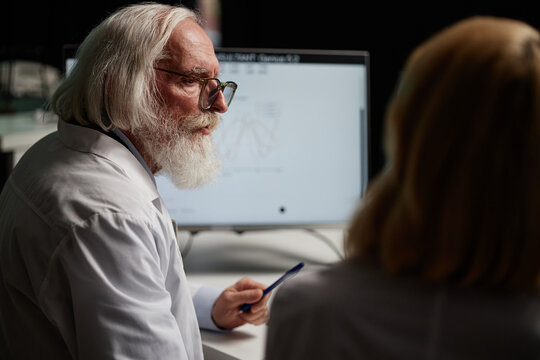 Senior Caucasian man with grey hair and beard discussing scientific data with middle aged woman in laboratory setting, both wearing lab coats, computer monitor displaying graph - Powered by Adobe