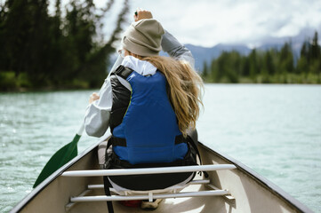 Back view of a young woman paddling a canoe across a clear, turquoise lake surrounded by pine trees...