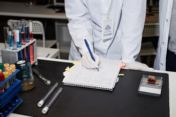 Unrecognizable Caucasian woman in lab coat writing scientific notes in notebook on laboratory table...
