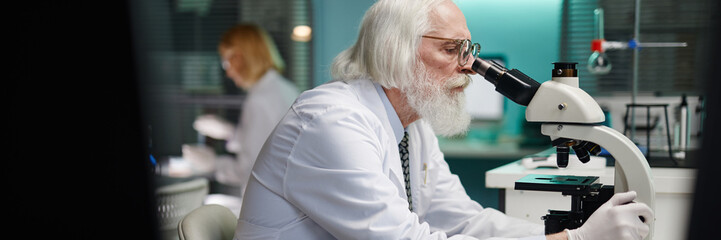 Website header shot of senior Caucasian man with grey hair and beard examining sample through microscope in scientific laboratory, female scientist working in background conducting research