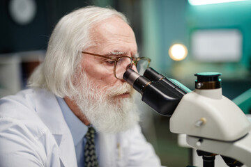 Senior Caucasian man examining specimen using laboratory microscope in scientific laboratory,...