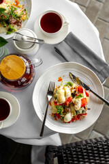 Top view of a fresh vegetable salad with creamy dressing, red tea, teapot, and sugar bowl on an elegant table setting with white tablecloth and napkin.