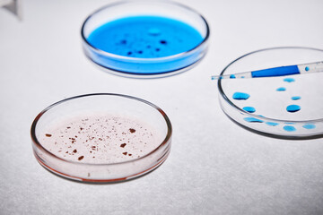 Close up showing three petri dishes with blue and pink liquid samples and pipette dispensing blue...